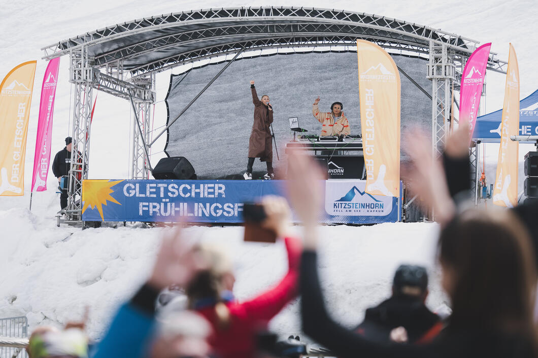 Konzert auf Bühne im Schnee, Sängerin mit erhobener Hand, DJ, jubelnde Menge