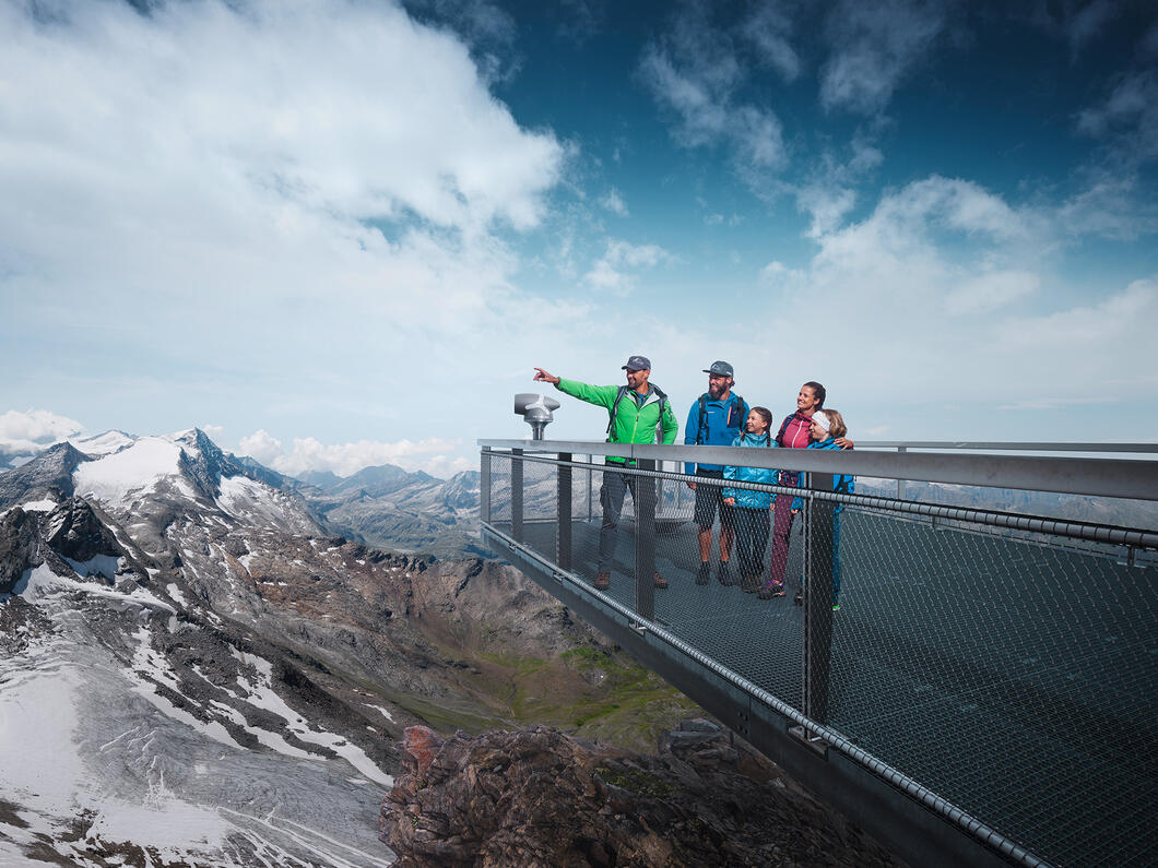Family on viewing platform in the mountains, man pointing into the distance, snow-capped peaks